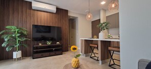Una sala de estar moderna con un televisor de pantalla plana, paneles de madera, una planta grande en maceta, una mesa redonda con un florero y una zona de bar con taburetes.