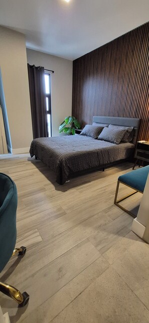 A modern bedroom with a large bed, a green armchair, a wooden floor, and a window with curtains.