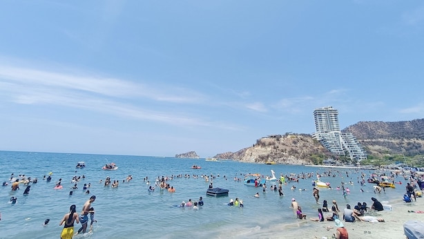 A crowded beach with people swimming and sunbathing, a large building in the background, and clear skies.