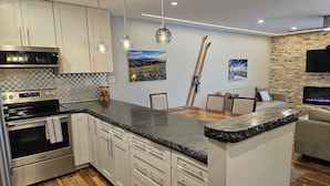A modern kitchen with a large island, stainless steel appliances, and a dining area with a stone wall and fireplace. - Center of Downtown Breck, walk to slopes and Main Street! Steps to Riverwalk. (Breckenridge)