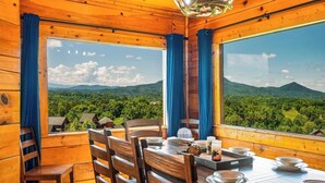 Un intérieur en bois avec une table à manger dressée pour quatre personnes, de grandes fenêtres offrant une vue sur un paysage montagneux.