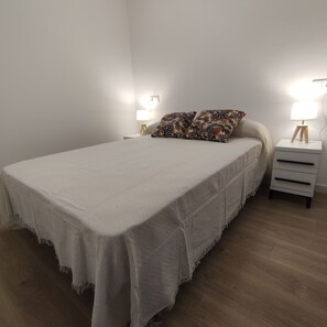 Un lit bien fait, recouvert d’une couvre-lit blanche et agrémenté de deux coussins décoratifs, est encadré par deux tables de chevet sur lesquelles reposent des lampes. La pièce est dotée d’un plancher en bois et de murs aux couleurs neutres.