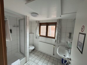 A bathroom with a shower, toilet, and sink, featuring white tiles and a window with curtains. - Hotel Sonne (Sinsheim)