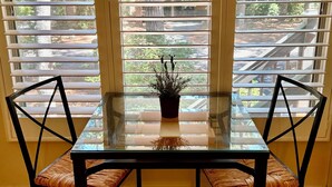 Un comedor con una mesa de vidrio y dos sillas, una planta en maceta sobre la mesa, y vistas a través de amplias ventanas con persianas.