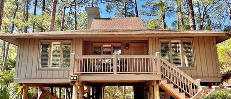 A wooden cabin with a balcony, elevated on stilts, surrounded by trees.