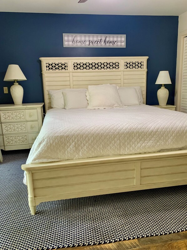 A bedroom with a white bed, a decorative headboard, two bedside tables with lamps, and a blue wall with a 'home sweet home' sign.