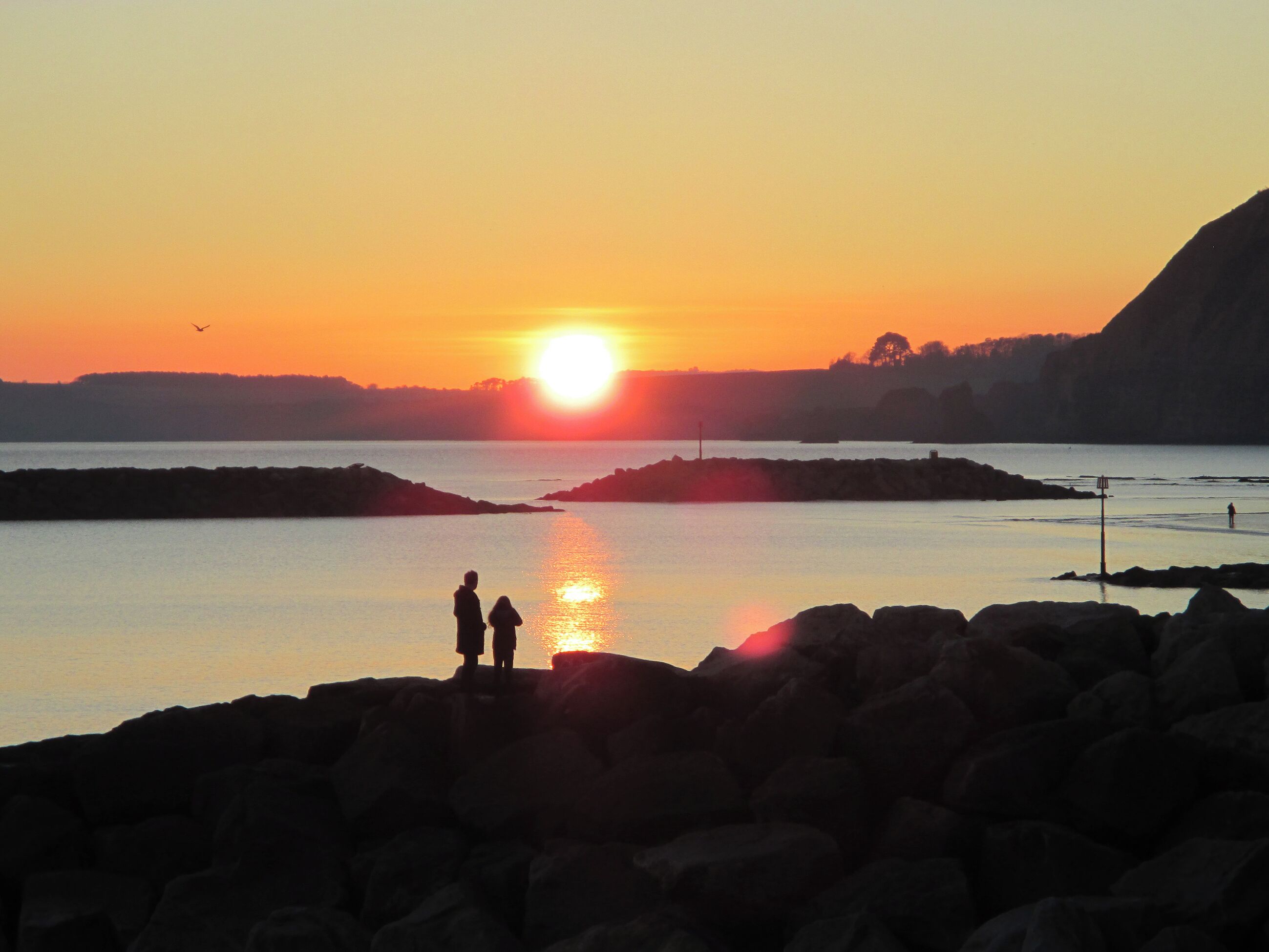 A sunset over a calm body of water with rocky shores and two silhouetted figures standing on the rocks.
