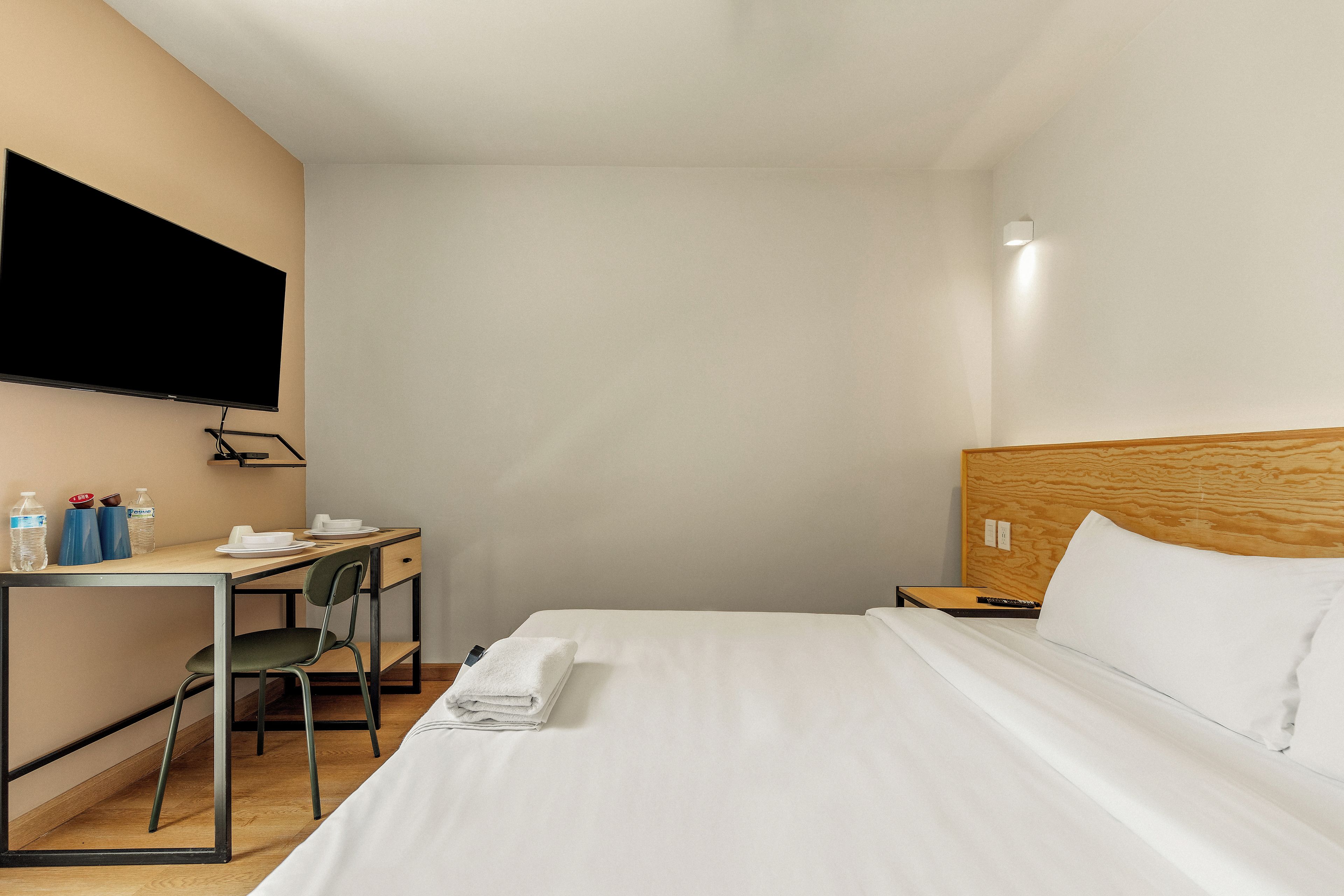 A hotel room with a bed, a desk, a chair, a TV mounted on the wall, and a wooden headboard.