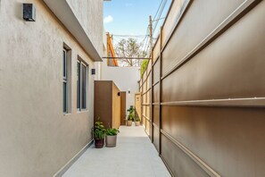 Ein schmaler Gang mit einem Gebäude auf einer Seite und einer Garage auf der anderen. Blumentöpfe und ein kleiner Baum sind zu sehen.