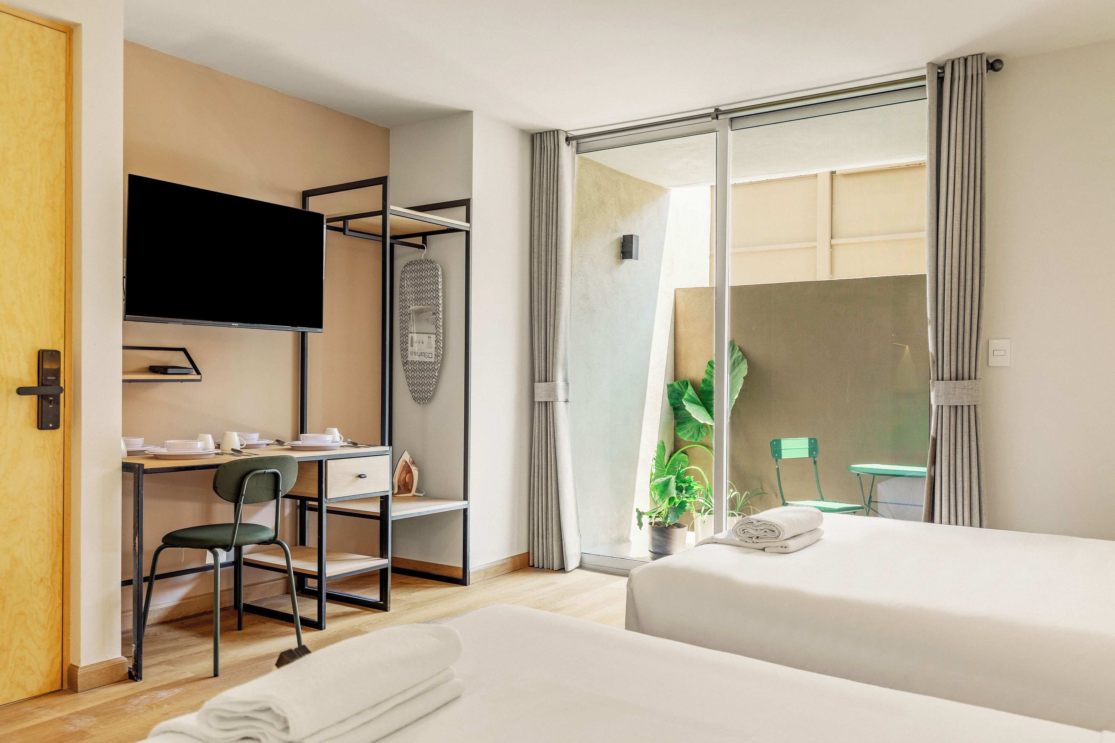 Habitación de hotel con escritorio, silla, televisión, dos camas, una planta en maceta y vistas a través de una puerta corredera.