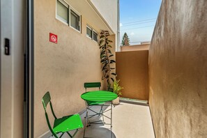 Um espaço ao ar livre estreito com mesa e cadeiras de metal verde, um vaso de planta e um edifício com parede bege e janelas.