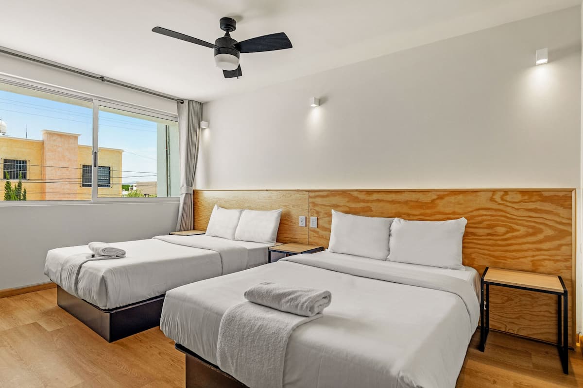A hotel room with two beds, a ceiling fan, a window with a view of buildings, and a wooden headboard.