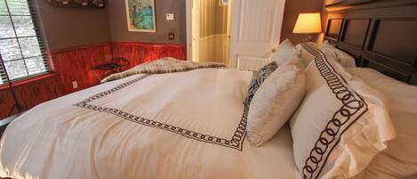 A neatly made bed with a decorative headboard, a red chair, and a painting on the wall.