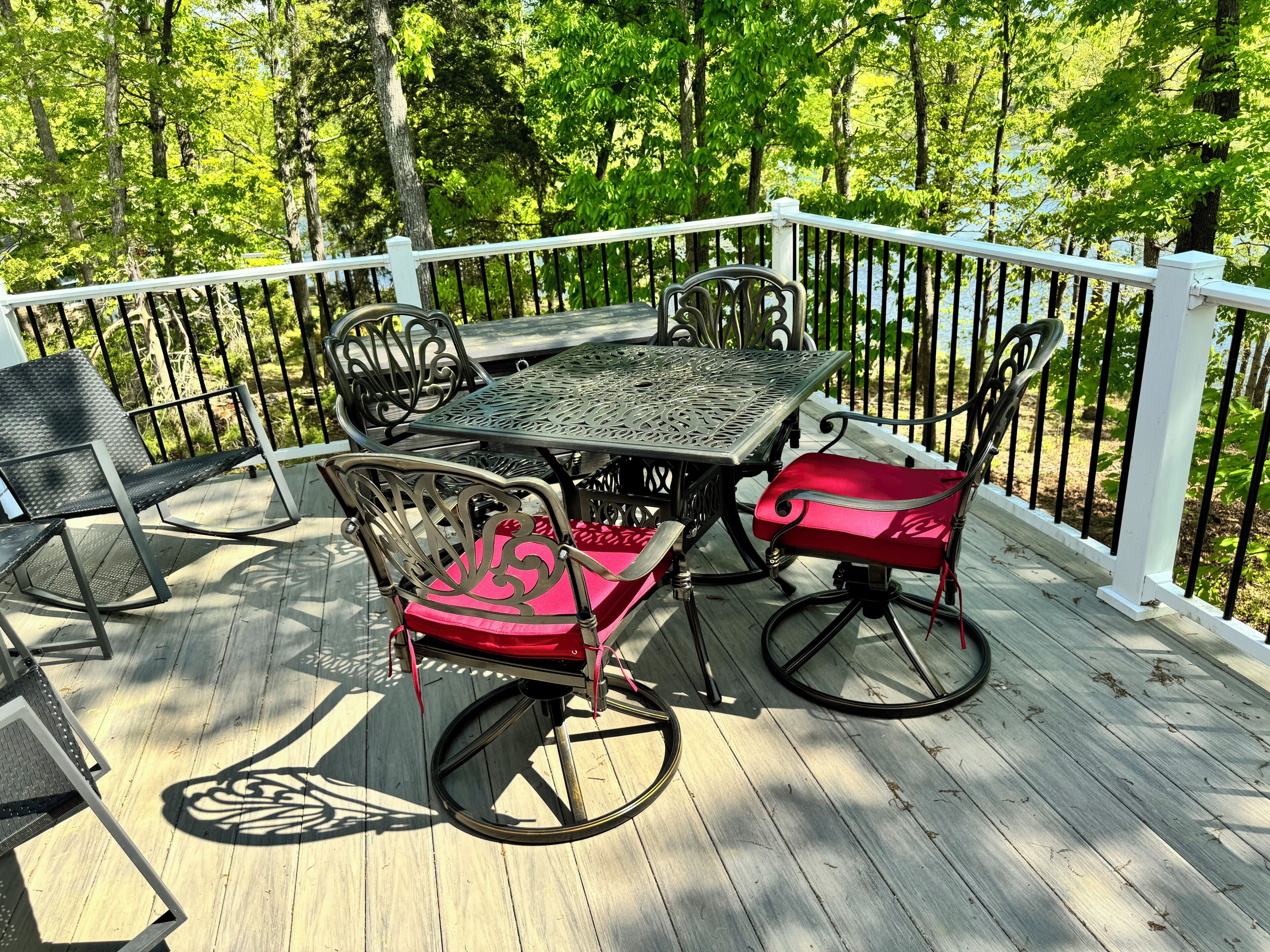 A wooden deck with a metal table and chairs, surrounded by a white railing and trees.