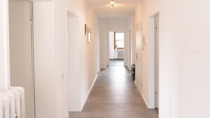 A long, narrow hallway with white walls and a light-colored floor, leading to a room with a radiator and a window.