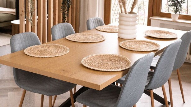 A dining area with a wooden table, grey upholstered chairs, woven placemats, and a hanging light fixture.