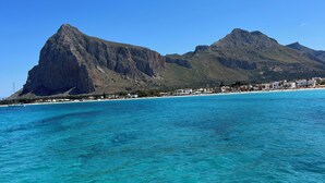 Escena costera con aguas turquesas cristalinas, una montaña rocosa en el fondo y una playa con edificios.