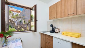 A compact kitchen with wooden cabinets, a coffee maker, a yellow tray, and a window with a view of a stone building. - A3 KABLAR'S SUNNY SUITES 1BR APT with balcony and sea view - Duplicate (Dubrovnik)