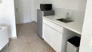 A compact kitchen with white cabinets, a microwave on top of a refrigerator, a sink, and a trash bin. - Rommar Cozy Suite (CAY HILL ,LITTLE BAY)