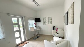 A modern, well-lit room with a glass table, white sofa, and a TV mounted on the wall. - Rommar Cozy Suite (CAY HILL ,LITTLE BAY)