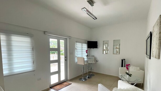 A spacious living room with a white sofa, a small table, a TV mounted on the wall, and a sliding glass door leading to a balcony. - Rommar Cozy Suite (CAY HILL ,LITTLE BAY)
