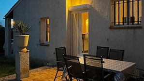 A white building with a patio area featuring a table and chairs, a potted plant, and a small window with bars.