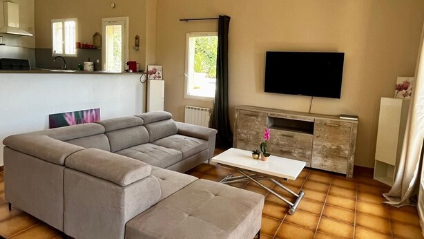 A living room with a sectional sofa, a flat-screen TV, a fireplace, and a kitchen area with a window.