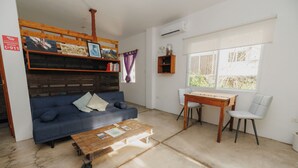 Ein Wohnzimmer mit einem blauen Sofa, einem hölzernen Couchtisch, einem kleinen Holztisch mit Stuhl und einem Fenster mit Blick auf Grünflächen.