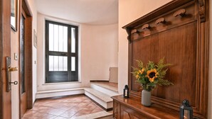 Ein Flur mit einer Holztür, einem Fenster mit Jalousien, einer Treppe und einem Konsolentisch mit Blumen und zwei Laternen.