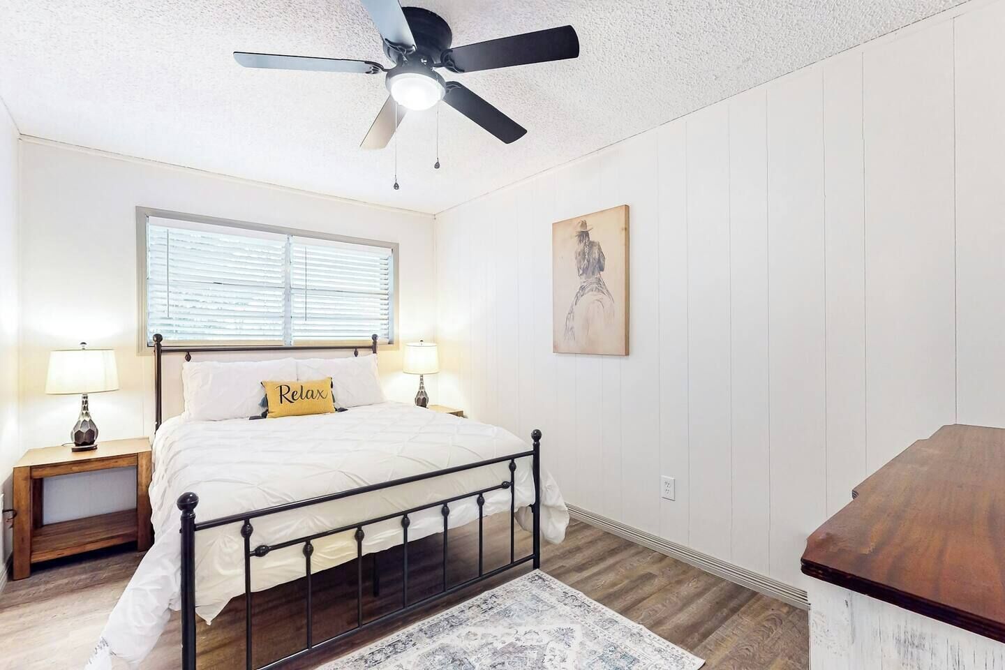 Un dormitorio con una cama, dos mesitas de noche, un ventilador de techo, una ventana con persianas, un cuadro en la pared y un aparador de madera.