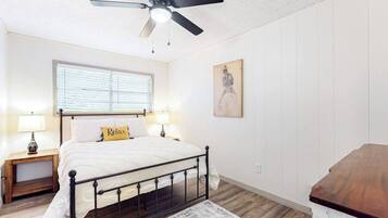 Un dormitorio con una cama, dos mesitas de noche, un ventilador de techo, una ventana con persianas, un cuadro en la pared y un aparador de madera.