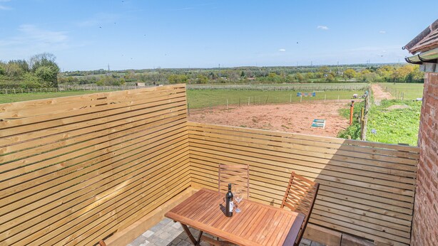 Une terrasse en bois avec une table et des chaises, d’où l’on peut admirer un champ verdoyant et des bâtiments situés au loin.