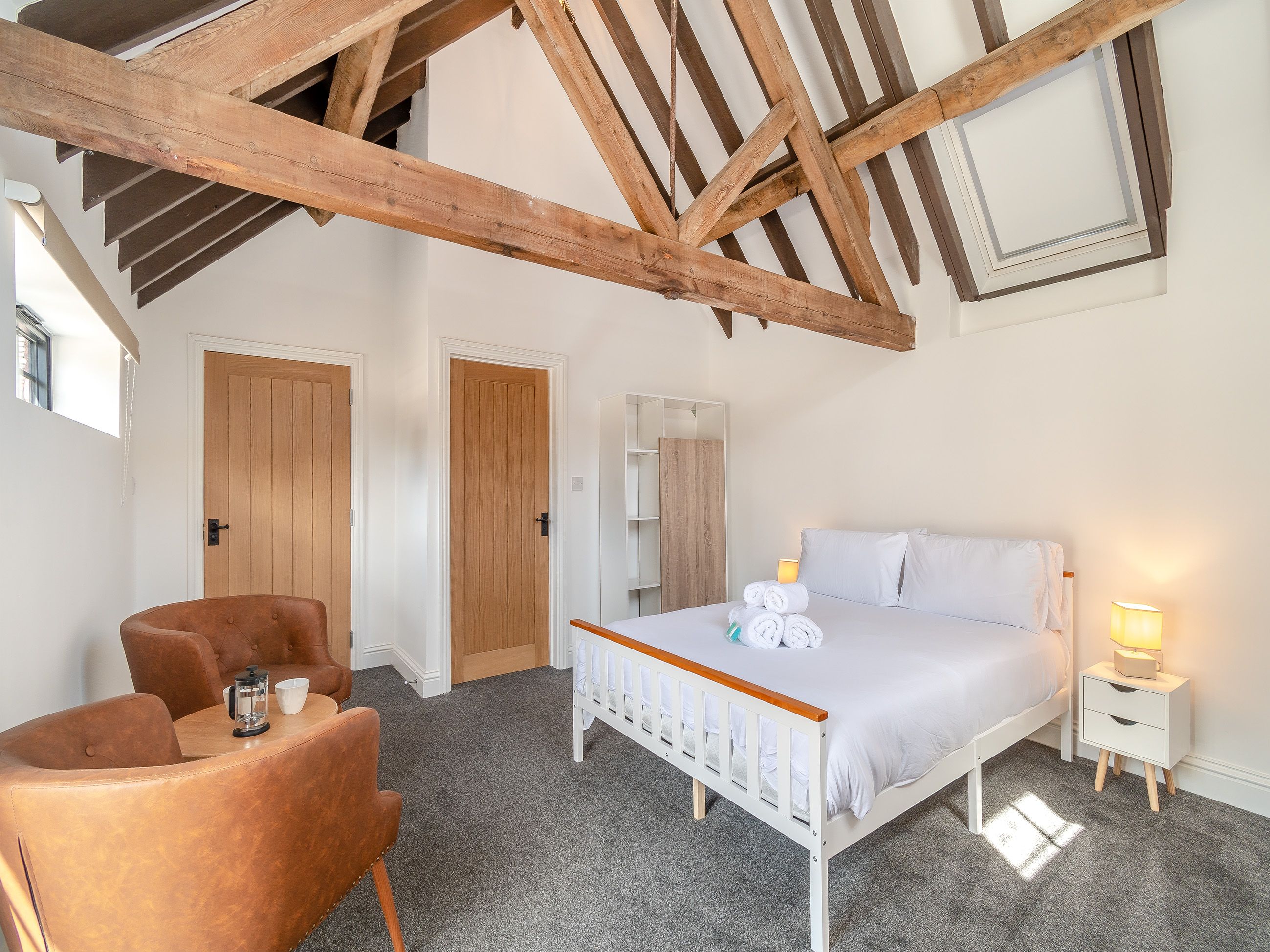 Un dormitorio con techo de madera, una cama con ropa de cama blanca, una mesita de noche con una lámpara y una silla con cafetera.