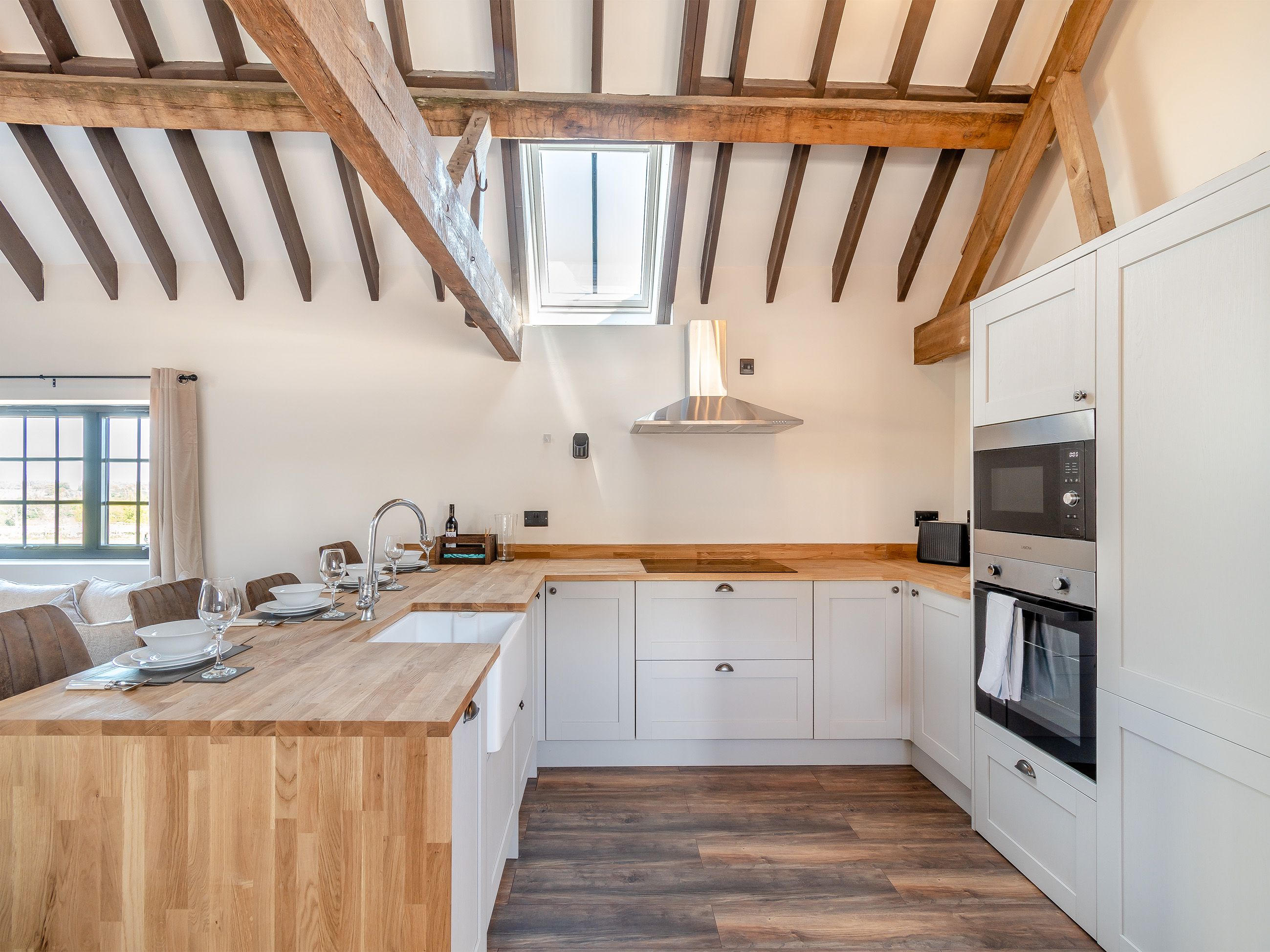 Una cocina moderna con vigas de madera, armarios blancos, horno empotrado y un ventanal grande.