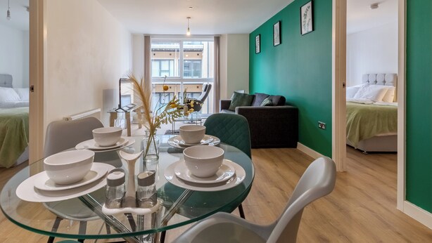 Een moderne woonkamer met een glazen eettafel voor vier personen, een groene bank en een bed met een wit hoofdeinde in de aangrenzende kamer.