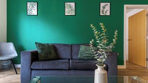 Een moderne woonkamer met een blauwgroene wand, een grijze bank met groene kussens, en een witte vaas met een plant.