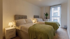 Een slaapkamer met twee bedden, een nachtkastje met een lamp, een kleine tafel met een plant en een raam met jaloezieën.