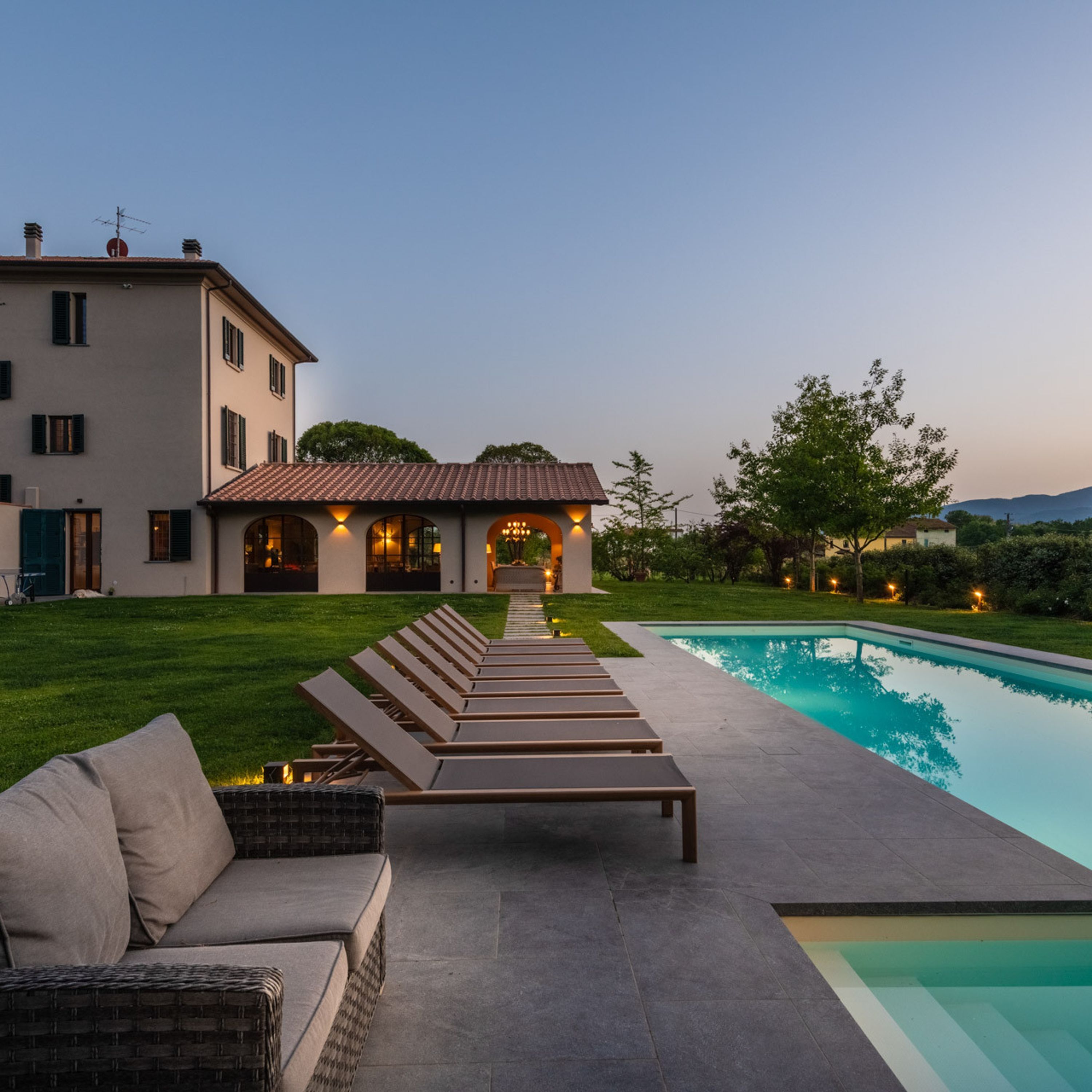 A poolside area with lounge chairs and a sofa, a swimming pool, and a building with a tiled roof and multiple windows.