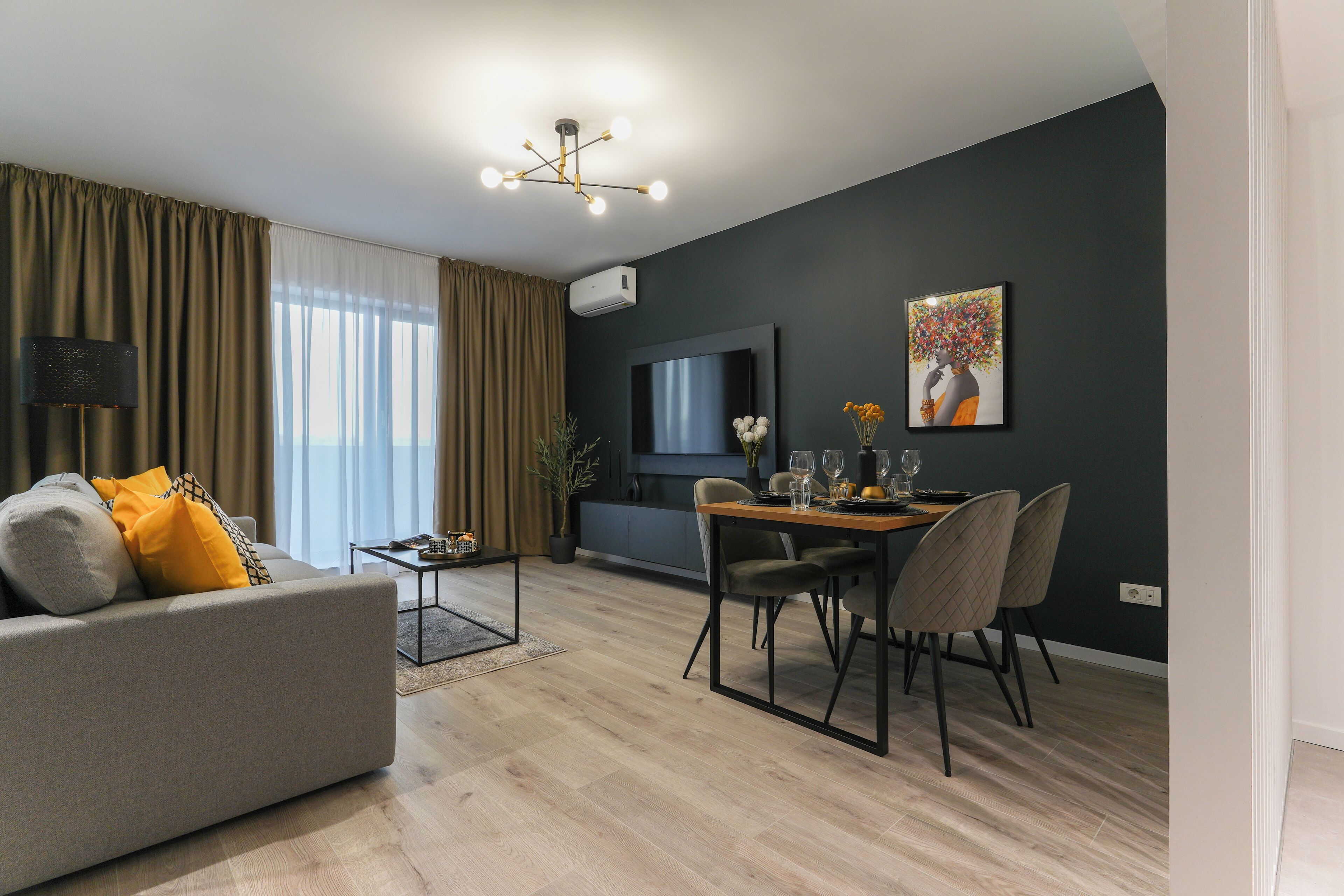 A modern living room with a sofa, dining table, and chairs. There is a flat-screen TV mounted on the wall and a potted plant on the windowsill.