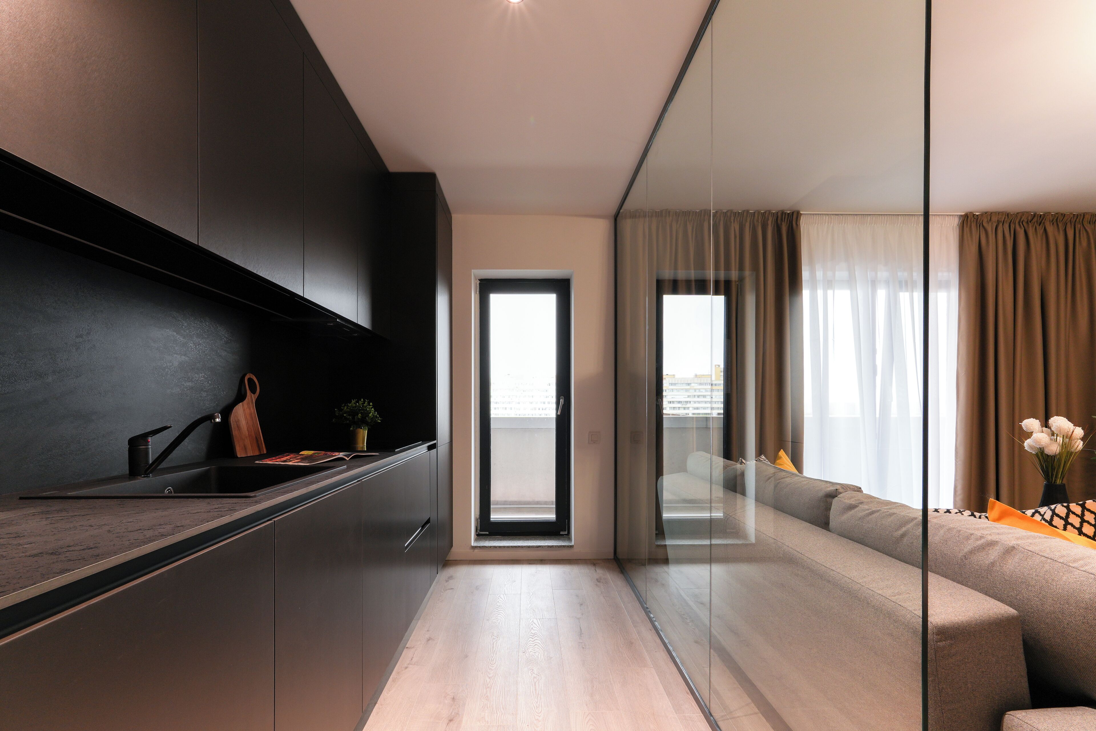A modern kitchen with a dark countertop, a sink, and a wooden cutting board. There is a sliding glass door leading to a living area with a sofa and a vase of flowers.