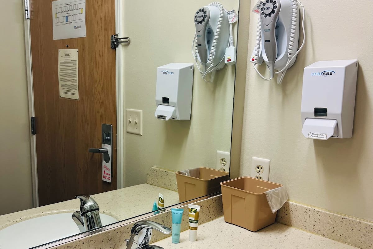 A hotel bathroom with a sink, mirror, hair dryers, and hand dryers.