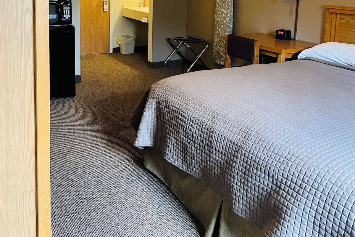 A hotel room with a bed, a desk, a chair, a microwave, a lamp, and a door.
