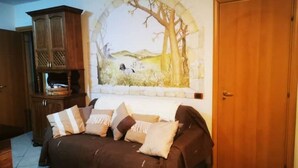 A cozy living room with a brown sofa, a wooden cabinet, and a mural of a tree and landscape on the wall. - 1 Bedroom Apartment in Procida (Procida)