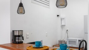 Ein Küchenbereich mit einer Kaffeemaschine, zwei blauen Tassen auf Untersetzern, einer Mikrowelle und einer weißen Wand mit Lüftung.