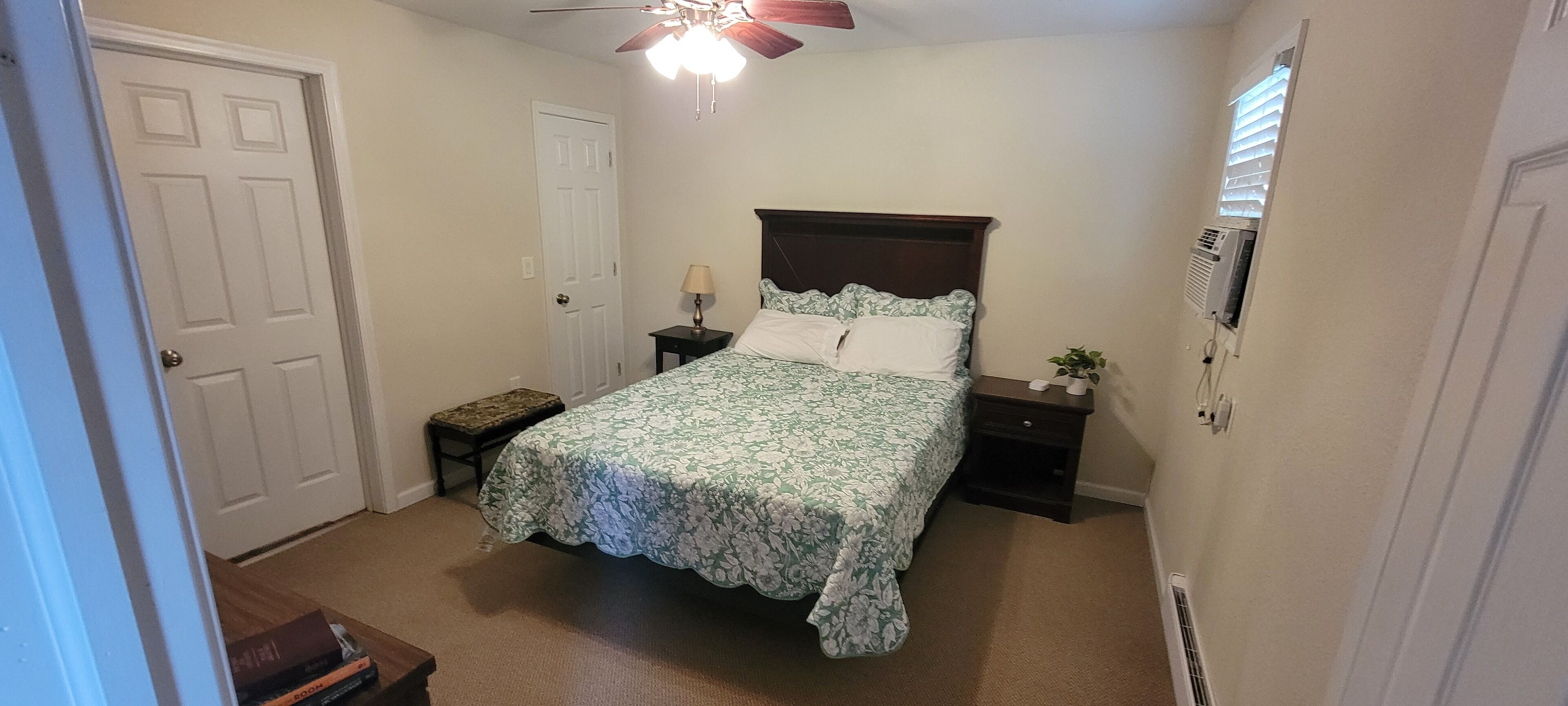 A bedroom with a bed, bedside tables, a bench, a ceiling fan, and a wall-mounted air conditioner.