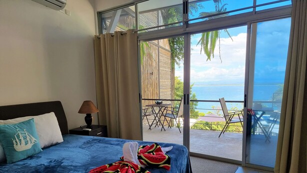 A bedroom with a bed, a nightstand, a lamp, and a view of an outdoor patio with a table and chairs. - S9 - Vista Ocean Suites (Quepos)