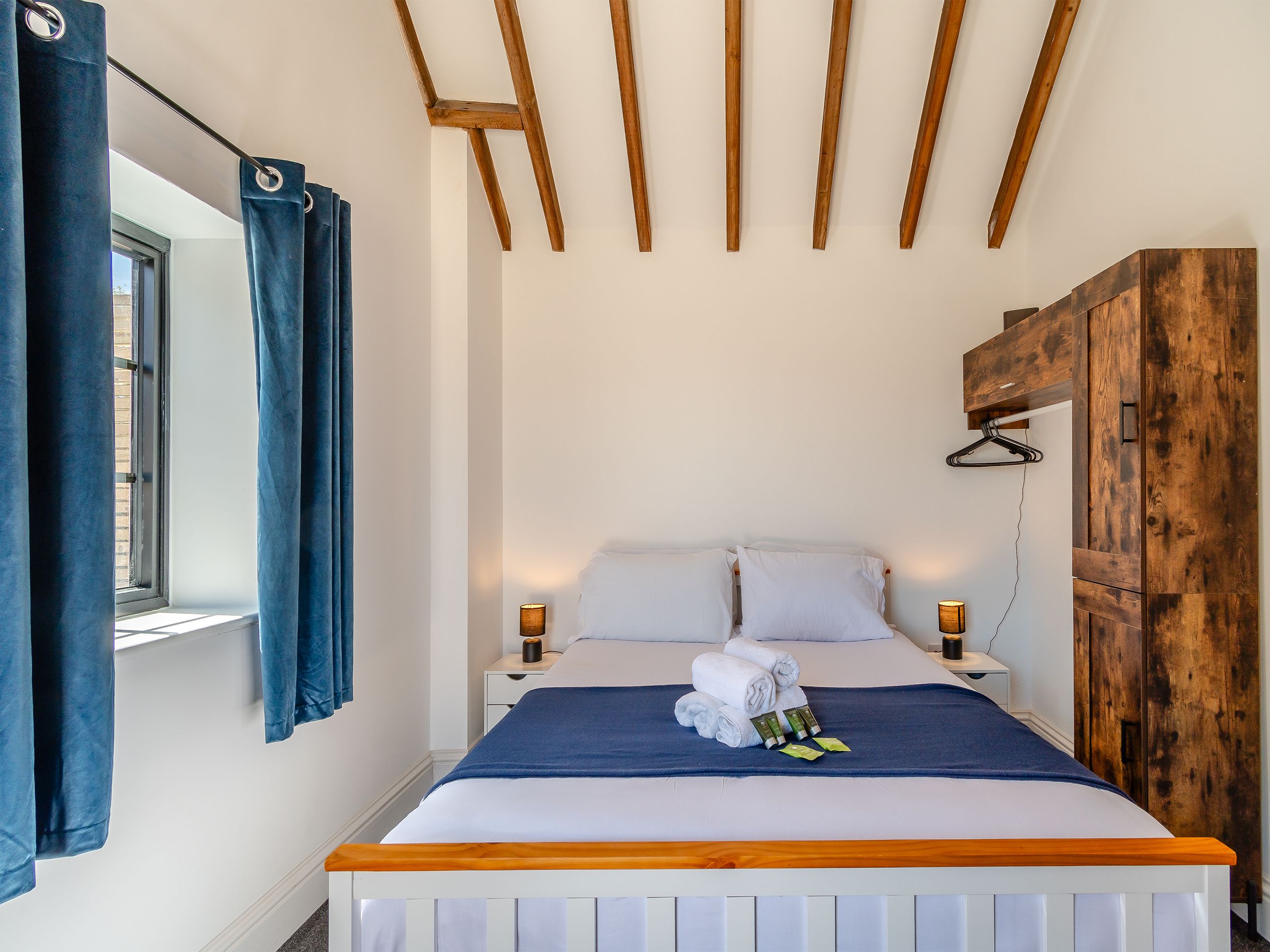 Ein ordentlich bezogenes Bett mit weißen Leinentüchern, einer blauen Decke und aufgerollten Handtüchern am Kopfteil. Es gibt zwei Nachttische mit Lampen, einen Holzschrank und ein Fenster mit blauen Vorhängen.