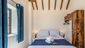 A neatly made bed with white linens, a blue blanket, and rolled towels on the headboard. There are two bedside tables with lamps, a wooden wardrobe, and a window with blue curtains. - Fountain Front Barn - UK45937 (Corley)