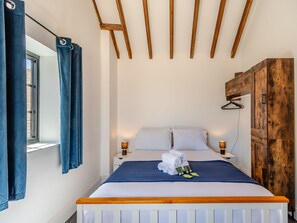 A neatly made bed with white linens, a blue blanket, and rolled towels on the headboard. There are two bedside tables with lamps, a wooden wardrobe, and a window with blue curtains. - Fountain Front Barn - UK45937 (Corley)