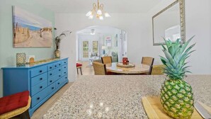 A kitchen with a granite countertop, a blue cabinet, a dining table with chairs, a pineapple on the counter, and a large mirror on the wall. - Ground Floor | Oceanfront Patio | Resort Style | P (Cape Canaveral)